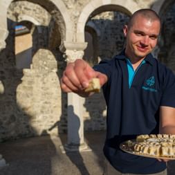 Reiseführer mit Inselkuchen in Kvarner Bucht Lächelnder Reiseführer in marineblauen Poloshirt hält Teller mit traditionellen Kuchenstücken vor Steinbögen in der Kvarner Bucht.