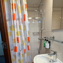 Kompaktes Badezimmer auf der Clair de Soleil mit weißen Fliesen, Waschbecken, Spiegel, Dusche mit buntem Fischvorhang und Toilettenartikeln.