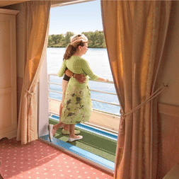 Frau in grünem Kleid auf privatem Balkon der Provence Suite mit Blick auf Fluss und bewaldetes Ufer. Zimmer mit beigem Teppich und Schrank.