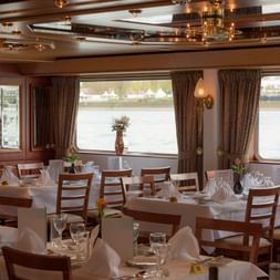 Elegant ship restaurant with white-clothed tables, wooden chairs, and large windows overlooking the water. Brown curtains and decorative ceiling.