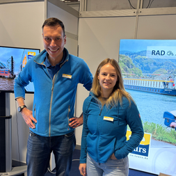 Zwei Mitarbeiter in blauen Jacken an einem Messestand mit Bildschirmen, die Flusskreuzfahrtschiffe und Radtouren zeigen.