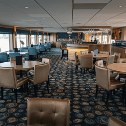 Geräumiges Restaurant auf der DE Nassau mit beigen Stühlen, runden Tischen, blau gemustertem Teppich und Barbereich. Große Fenster sorgen für Tageslicht.