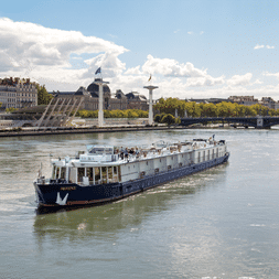 Flusskreuzfahrtschiff Provence auf der Rhône mit blauem Rumpf und weißem Aufbau. Französische Flaggen sichtbar, Stadtufer mit Gebäuden im Hintergrund.