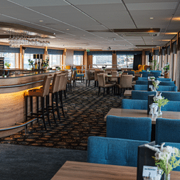 Moderne Lounge auf der MS Nassauer mit geschwungener Holzbar, blau gepolsterten Sitzen und großen Fenstern mit Blick aufs Wasser.