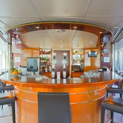 Interior of MS SE-MANON salon featuring a circular wooden bar with bar stools, purple carpeting, and large windows providing natural light.