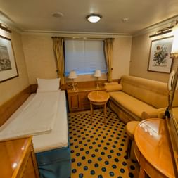 Innenraum der MS Casanova Hauptdeck-Kabine mit zwei Einzelbetten, gelbem Sofa, rundem Tisch, Fenster mit Vorhängen und blau gemustertem Teppich.