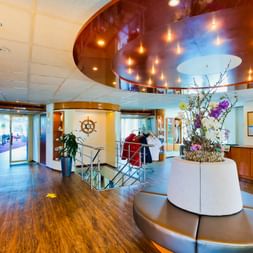 Modern ship reception area on MS SE-MANON with curved ceiling lights, wooden floors, circular seating, and nautical wheel decoration.
