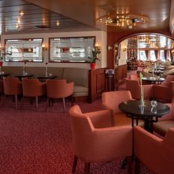 Elegant salon aboard the Switzerland ship with coral-colored armchairs, round tables, red carpet, and wood-paneled ceiling with chandeliers.