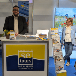Zwei Personen am se-tours Stand: Mann im schwarzen Anzug hinter Theke, Frau in blauer Jacke neben Prospektständer. Fahrradrad und Landschaftsbild sichtbar.