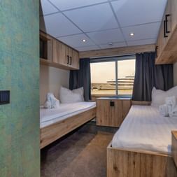 Modern upper deck cabin on MS PRINCESS with two single beds, wood furnishings, large window, and blue curtains. White towels arranged on beds.