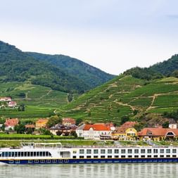 MS SE-MANON river cruise ship sailing on calm waters with terraced vineyards on hillsides and colorful village houses in background.