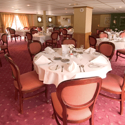 Eleganter Speisesaal auf dem Schiff Provence mit runden Tischen mit weißen Tischdecken, burgunderroten Polsterstühlen und rosa gemustertem Teppich.