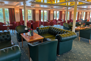 Eleganter Salon an Bord der Casanova mit grünen Chesterfield-Ledersofas, roten Samtstühlen, Holztischen, weißen Säulen und großen Fenstern.