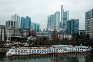 Weißes Flusskreuzfahrtschiff MS Classica mit orangefarbenem Streifen auf Fluss vor moderner Skyline mit Hochhäusern.