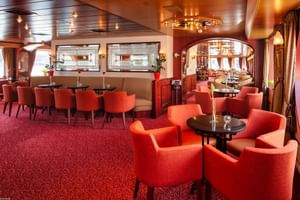 Elegant salon bar on MS Switzerland with red carpeting, orange upholstered chairs, round tables, and decorative mirrors on walls.