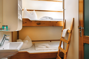 Compact ship cabin on Mare Frisium with wooden bunk beds, white linens, towels, wooden ladder, sink, and storage shelves.