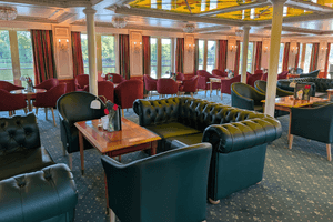 Elegant salon aboard the Casanova ship with green leather Chesterfield sofas, red velvet chairs, wooden tables, white columns, and large windows.