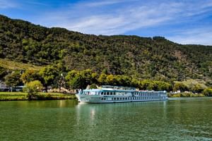 White river cruise ship MS Switzerland sailing on calm green water with forested hills and village buildings in the background under blue sky.