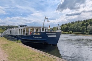 River cruise ship Casanova with dark blue hull and white upper deck moored at grassy riverbank under cloudy sky with green trees in background.