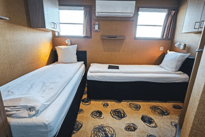 Compact twin cabin on DE Nassau with two single beds, white bedding, two windows with curtains, air conditioning unit, and patterned carpet.