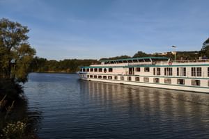 Weißes Flusskreuzfahrtschiff Switzerland mit grünem Zierstreifen auf ruhigem Fluss. Bäume säumen das linke Ufer, Gebäude auf fernen Hügeln.