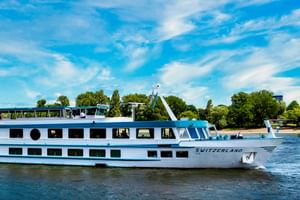 White river cruise ship MS Switzerland sailing on calm water with blue sky, trees and greenery visible along the shore in the background.