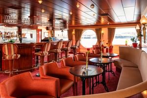 Elegant salon bar on MS Switzerland with orange and beige seating, dark wood paneling, round porthole windows, and a bar counter with glassware.