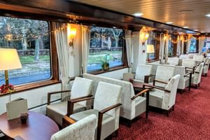 Innenraum des MS Classica Salons mit Reihen weißer Sessel, rotem Teppich, großen Fenstern mit Vorhängen und warmer Beleuchtung. Bäume draußen sichtbar.