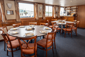 Speisesaal auf dem Schiff Clair de Soleil mit runden und rechteckigen Tischen, Holzstühlen, Panoramafenstern und Barbereich.