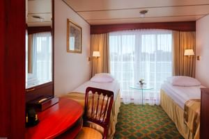 Twin cabin on MS Switzerland with two single beds, large windows with curtains, green patterned carpet, and wooden furniture including desk and chair.