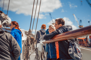 Passengers on the deck of sailing ship Mare Frisium enjoying a sunny day. People in casual jackets lean against wooden railings with rigging visible.