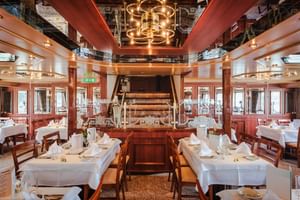 Elegant restaurant aboard MS Switzerland with white-clothed tables, wooden paddle wheel centerpiece, and brass chandelier under mirrored ceiling.