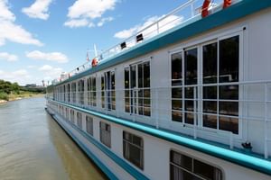 Weißes Flusskreuzfahrtschiff mit türkisfarbenen Akzenten und großen französischen Balkonfenstern am Fluss unter blauem Himmel mit Wolken.