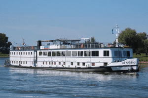 White river cruise ship De Nassau sailing on calm water. The multi-deck vessel has large windows and a sun deck, with green riverbanks visible.
