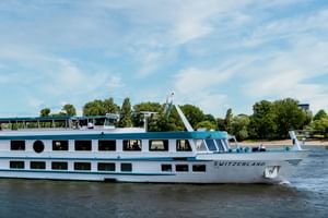 Weißes Flusskreuzfahrtschiff namens Switzerland fährt auf einem Fluss mit grünen Bäumen und blauem Himmel im Hintergrund. Das Schiff hat zwei Decks mit Fenstern.