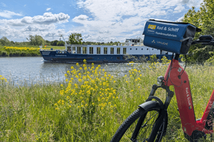 River cruise ship Casanova with blue and white hull on waterway. Red bicycle with tour company sign in foreground, yellow wildflowers along shore.
