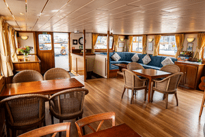 Heller Salon an Bord der Clair de Soleil mit Holzboden, Korbstühlen, blauem Sofa, goldenen Vorhängen und großen Fenstern mit Blick auf das Deck.