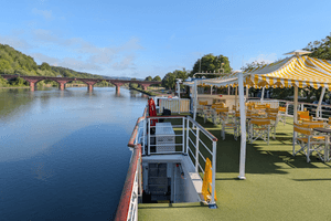 Sun deck of river cruise ship Casanova with yellow-white striped awning, wooden chairs, and green artificial turf. River and bridge visible in background.