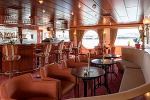 Elegante Salon Bar auf dem Schiff Switzerland mit Holztheke, Ledersesseln, runden Tischen und runden Bullaugen mit Blick aufs Wasser.