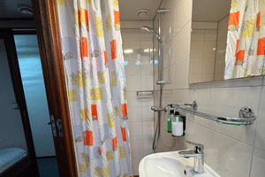 Kompaktes Badezimmer auf der Clair de Soleil mit weißen Fliesen, Waschbecken, Spiegel, Dusche mit buntem Fischvorhang und Toilettenartikeln.