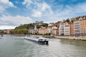 Flusskreuzfahrtschiff Provence auf einem Fluss mit bunten historischen Gebäuden am Ufer und einer Hügelstadt unter blauem Himmel.