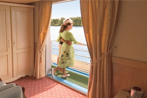 Frau in grünem Kleid auf privatem Balkon der Provence Suite mit Blick auf Fluss und bewaldetes Ufer. Zimmer mit beigem Teppich und Schrank.