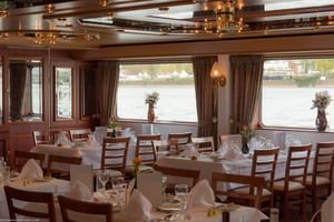 Elegantes Schiffsrestaurant mit weiß gedeckten Tischen, Holzstühlen und großen Fenstern mit Blick aufs Wasser. Braune Vorhänge und dekorative Decke.
