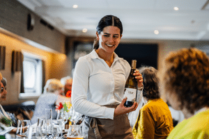 Smiling staff member in white polo shirt presenting wine bottle to guests in the Magnifique X restaurant dining area.