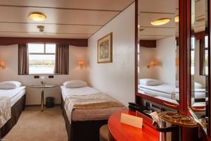 Twin cabin on MS Switzerland main deck with two beds, window view, vanity desk, mirror, wall lamps, and beige-brown decor.