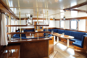 Interior lounge of Mare Frisium ship with blue upholstered benches, wooden tables, brass poles, polished wooden floors, and white ceiling.