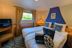 Ship cabin on Casanova with two twin beds, blue accent wall, window with curtains, TV, and cycling equipment. White bedding and patterned carpet.