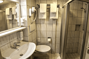 Kompaktes Badezimmer auf MS Classica mit beigen Fliesen, weißem Waschbecken, WC, Glasduschkabine, Wandhaartrockner und weißen Handtüchern.