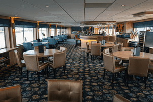 Spacious restaurant on DE Nassau with beige chairs, round tables, blue patterned carpet, and bar area. Large windows provide natural light.