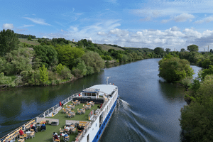 Luftaufnahme des Flusskreuzfahrtschiffs Casanova mit Sonnendeck und Passagieren in grüner Landschaft mit üppigen Bäumen unter blauem Himmel.